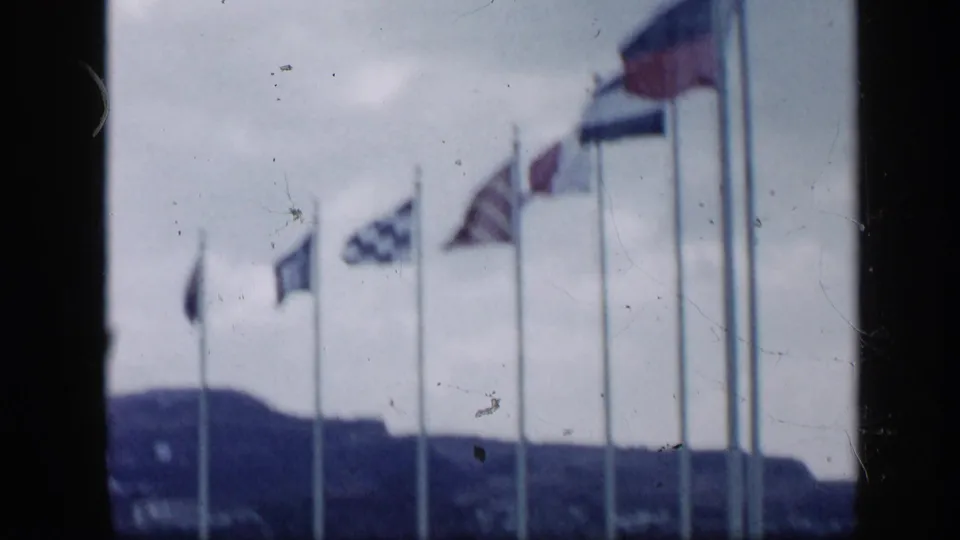 1959 Venezuela home-movie frame with a row of flags on tall poles blowing in wind under gray sky, with hills beyond.