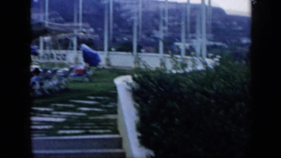 1959 Venezuela home-movie frame showing steps, clipped hedges, flowers, and a glassy garden structure under overcast light.