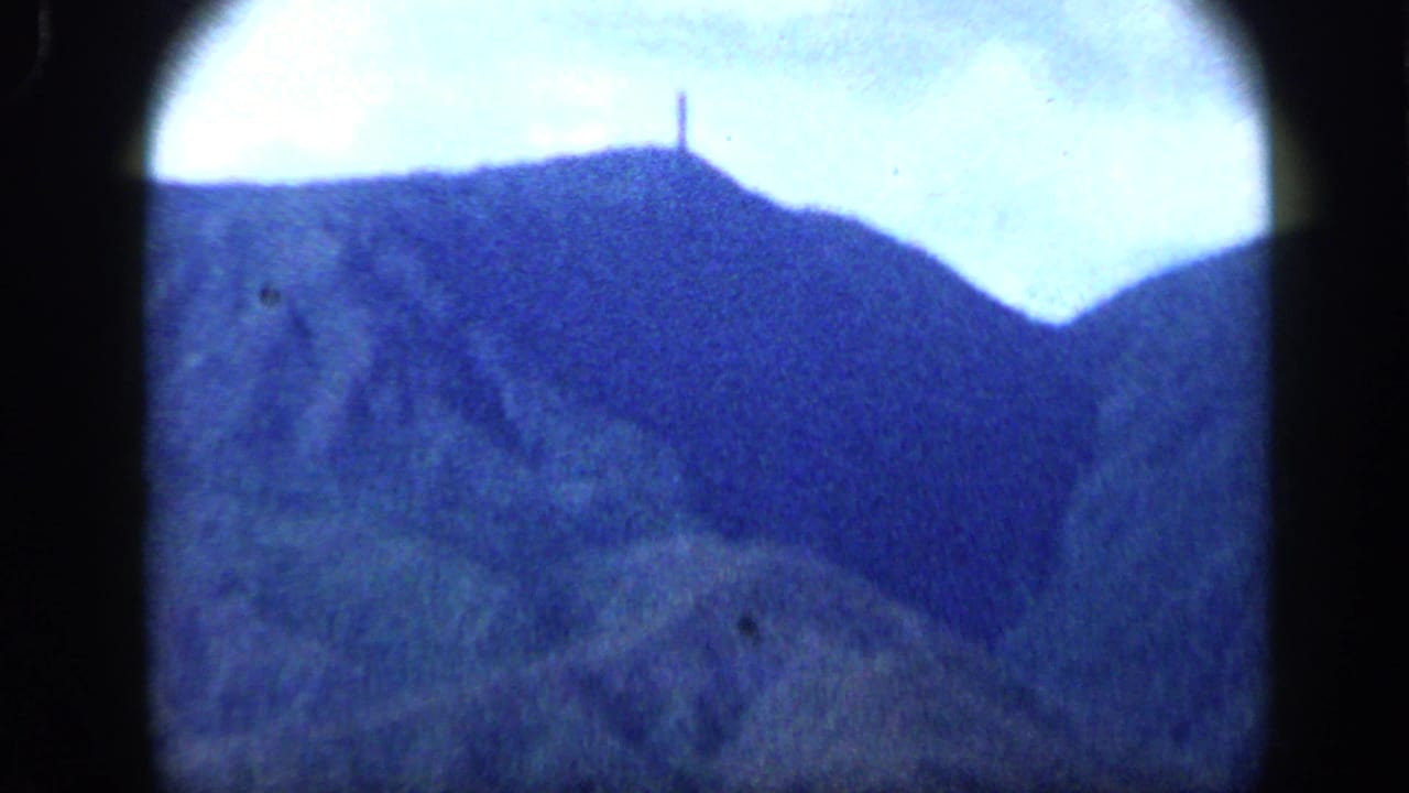 Grainy 1959 8mm frame from Venezuela showing a mountain ridge and a small vertical monument silhouetted near the top.