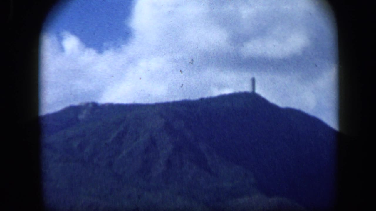 1959 Venezuela 8mm frame showing a cloudy mountain skyline, a small summit statue, and a tiny aircraft overhead.