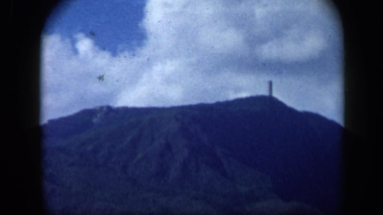 1959 Venezuela 8mm frame showing a dark mountain ridge below a cloudy sky, with a slim structure on the hilltop.