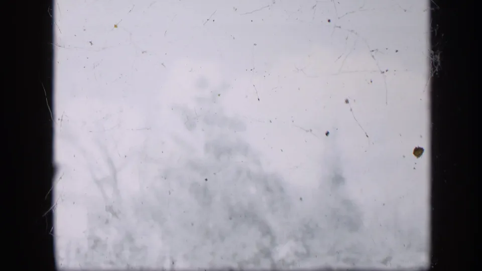 Pale 8mm frame with visible scratches, dust, and faint out-of-focus forms during a damaged or exposed section of the clip.