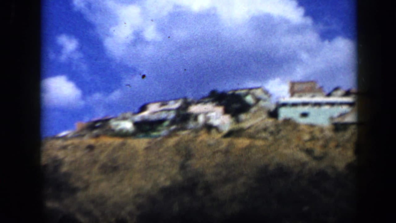 Daytime 1959 Venezuela home-movie frame showing houses on a dry hillside beneath a bright blue, cloudy sky.