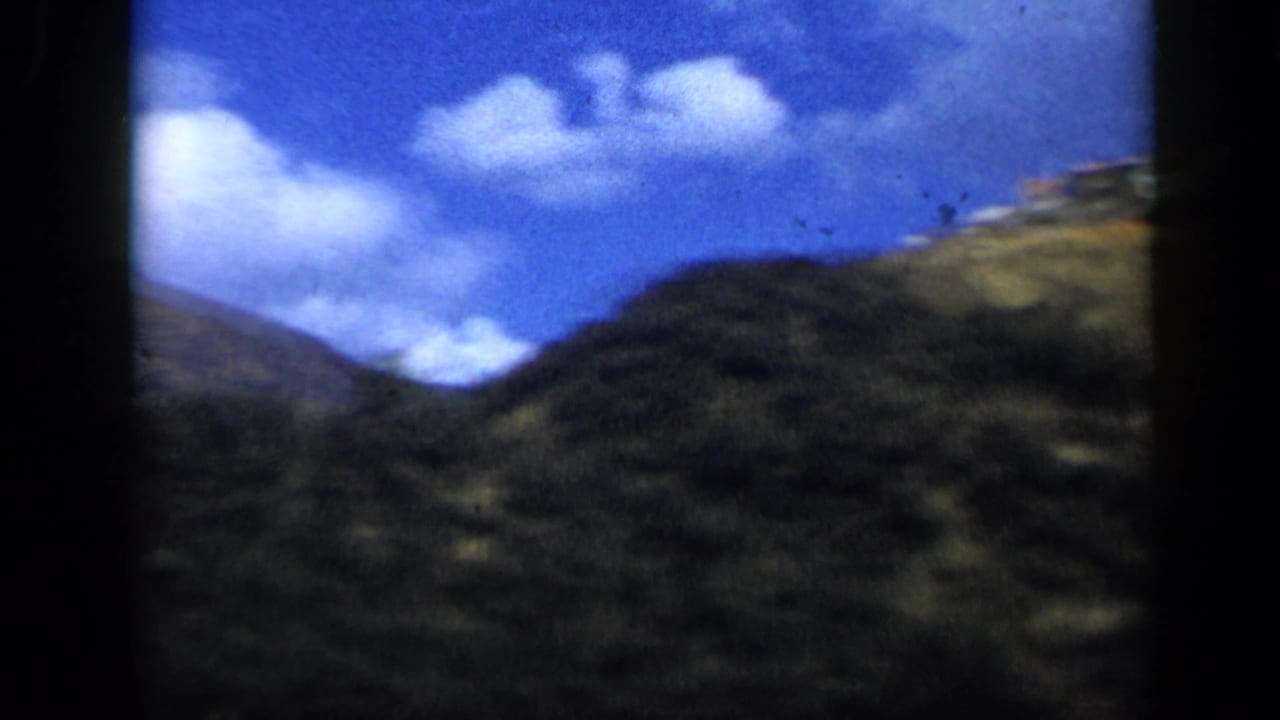 1959 Venezuela 8mm frame showing a bright mountain hillside, blue sky, and scattered clouds seen from a moving countryside drive.