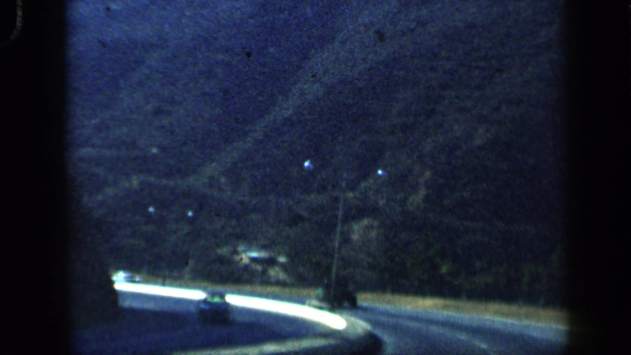 1959 Venezuela countryside road curves beside a white barrier, with bare trees and dark mountain slopes in the background.