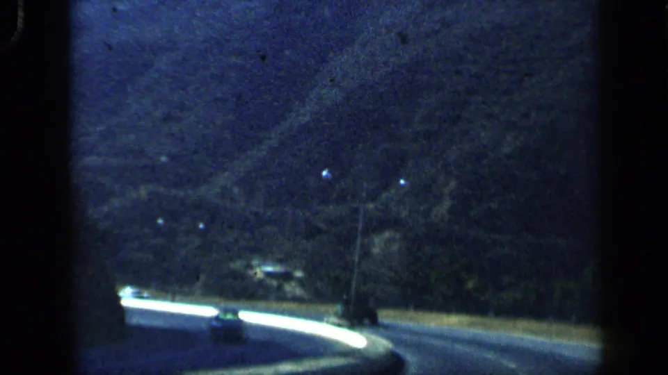 1959 Venezuela countryside road curves beside a white barrier, with bare trees and dark mountain slopes in the background.