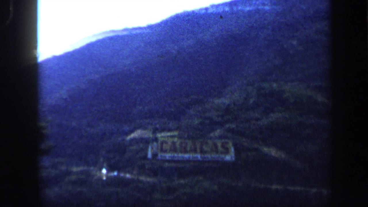 1959 Venezuela home-movie frame showing a distant Caracas sign set into a wooded hillside beneath blue-toned mountains.