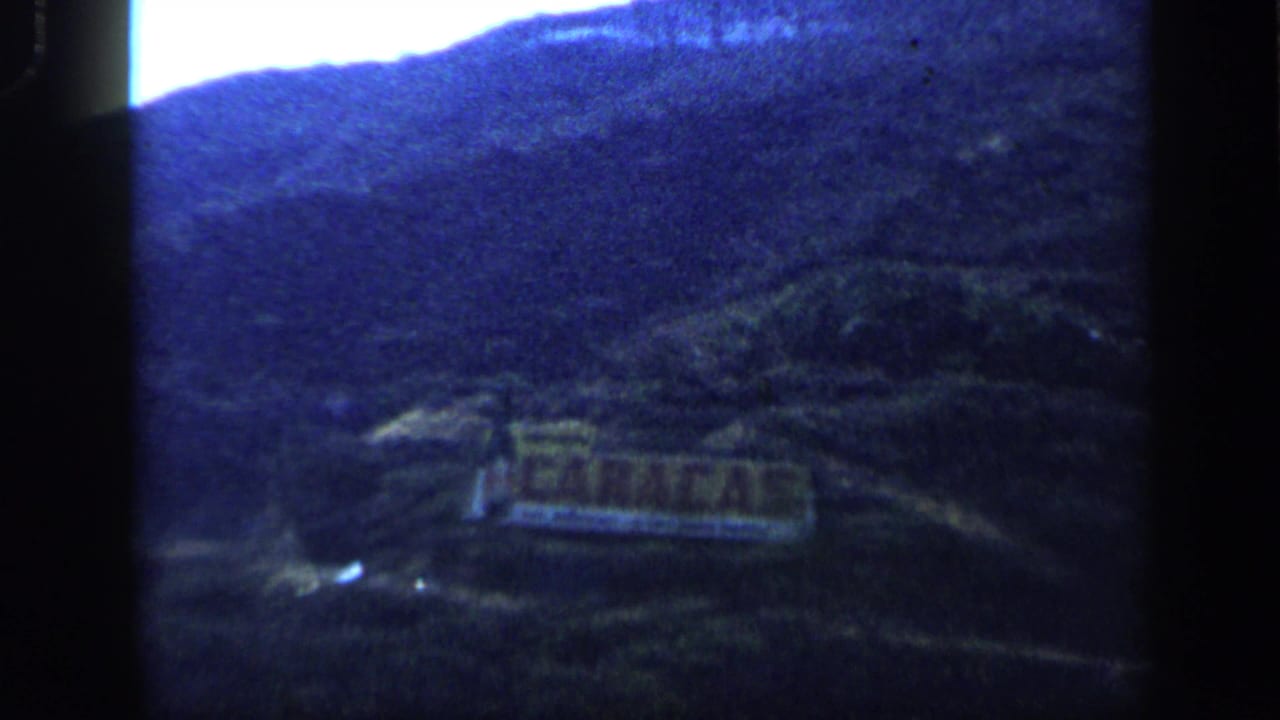 Blue-tinted 8mm frame of the Caracas hillside sign, with mountains rising behind and dark film-frame borders visible.