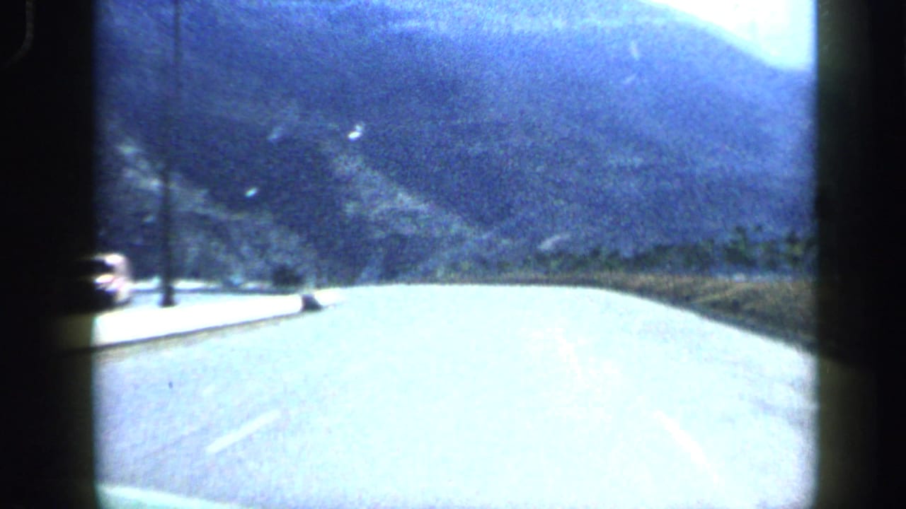 Visitor-shot 1959 road view in Venezuela, with a pale paved roadway, roadside poles, trees, and hazy mountains ahead.