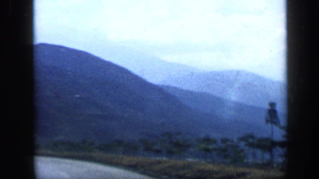 Blue-toned 8mm view from the road toward Venezuelan mountains, with distant low buildings and a small roadside signpost.