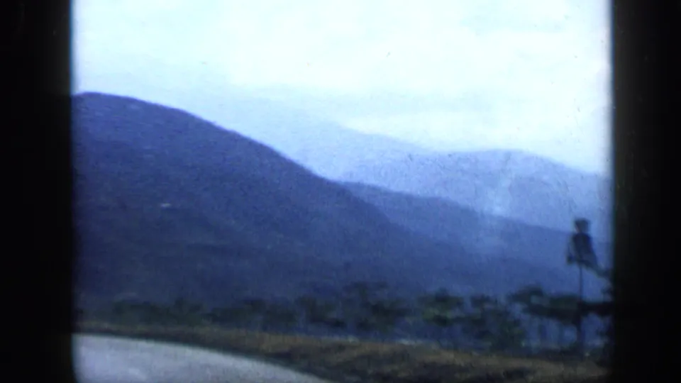 Blue-toned 8mm view from the road toward Venezuelan mountains, with distant low buildings and a small roadside signpost.
