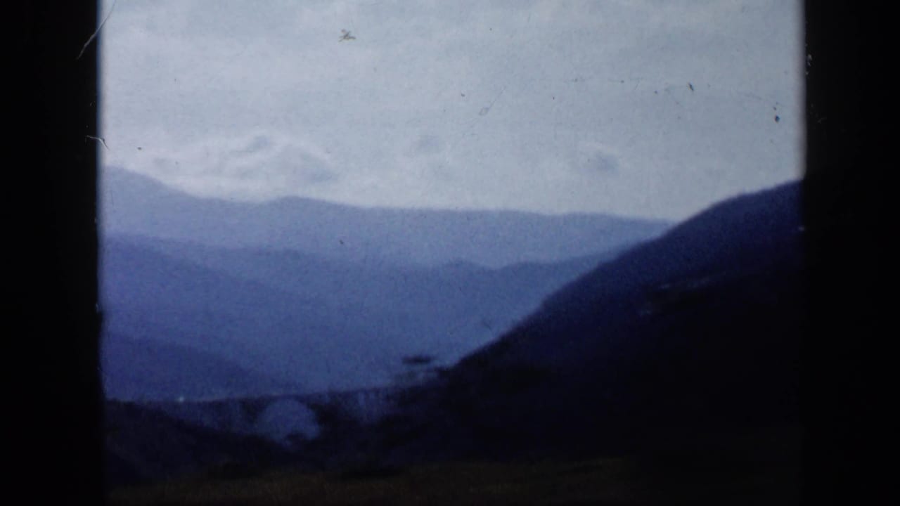 1959 home-movie view of a Venezuelan mountain valley, with a dark hillside and a faint bridge or roadside structure below.