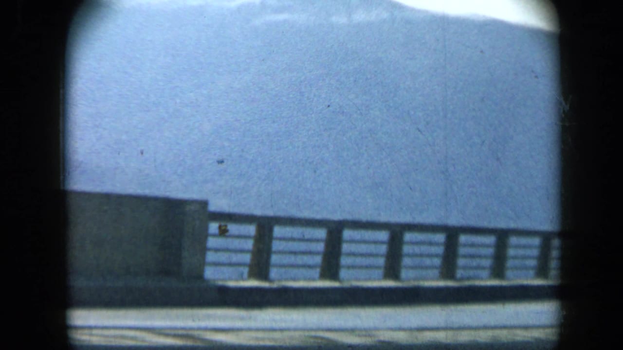 A roadside bridge or barrier passes the window, with a pale sky and distant hillside visible in the 1959 Venezuela footage.