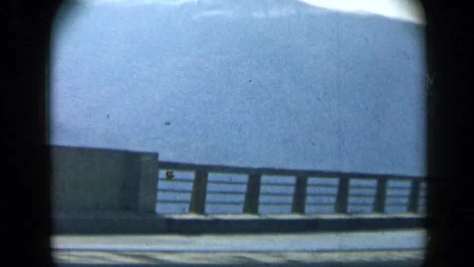 A roadside bridge or barrier passes the window, with a pale sky and distant hillside visible in the 1959 Venezuela footage.
