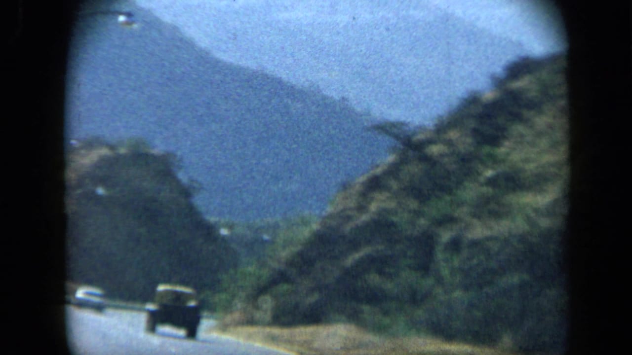 1959 Venezuela mountain road scene with a dark vehicle ahead and layered hills in the distance.