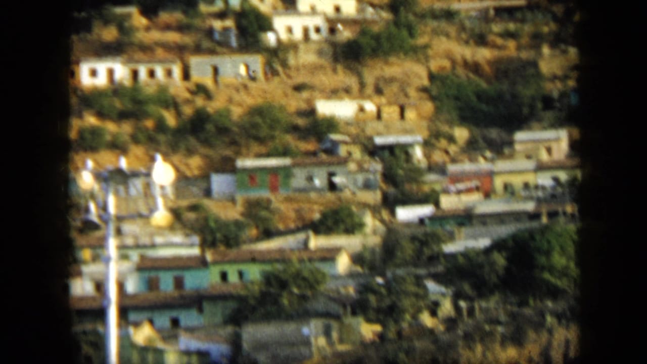 1959 Venezuela 8mm frame showing small hillside houses, a utility pole, and vegetation beneath a mountain slope.