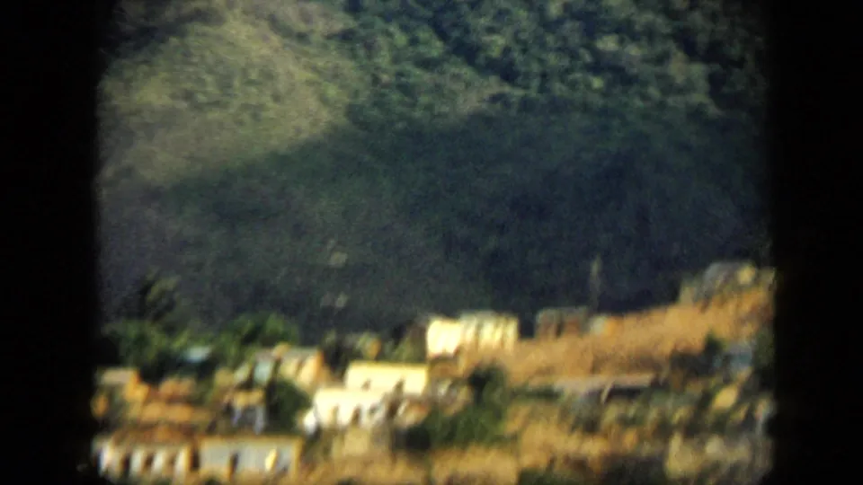 Soft 8mm view of a Venezuelan hillside settlement with pale buildings, dry ground, and dark green mountain vegetation.