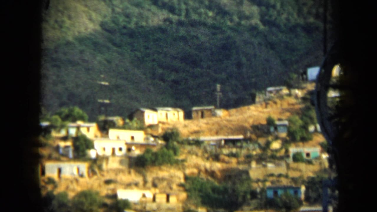 Vintage Venezuela frame with small hillside houses, scrubby slopes, utility lines, and dense green vegetation above.