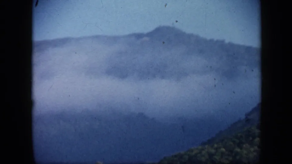 1959 Venezuela 8mm view of a hazy mountain ridge with mist or smoke across the slope and visible film dust.