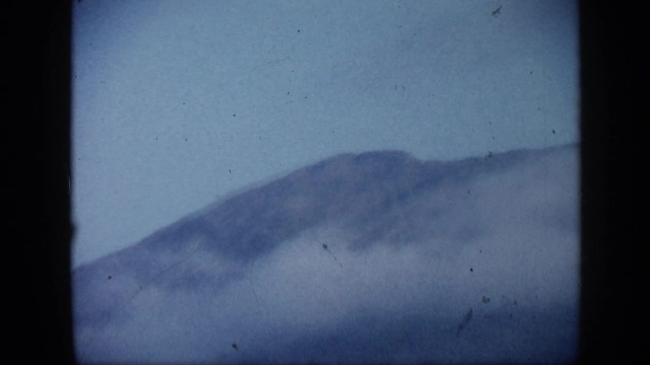 1959 Venezuela 8mm landscape frame with a softened mountain ridge, pale sky, and mist or smoke covering the lower view.