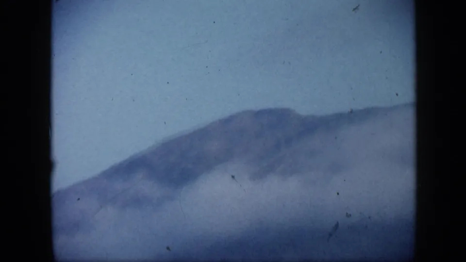 1959 Venezuela 8mm landscape frame with a softened mountain ridge, pale sky, and mist or smoke covering the lower view.