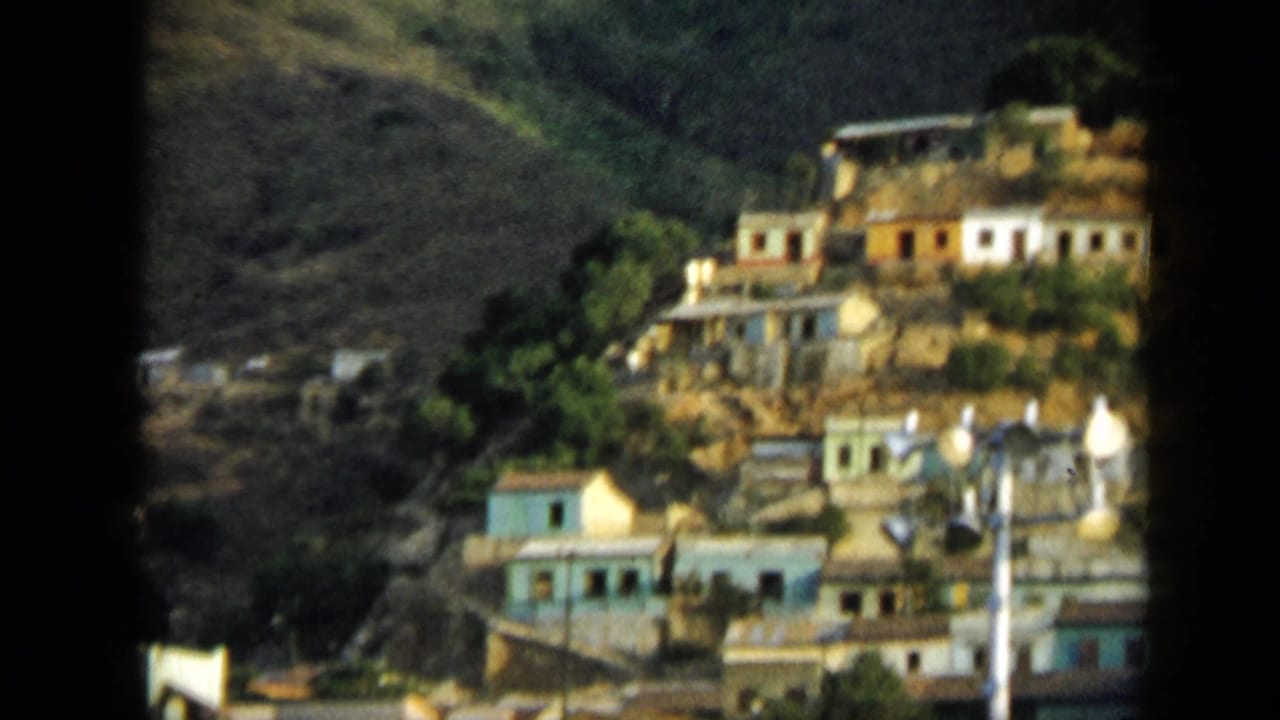 1959 Venezuela home-movie frame showing small colorful houses clustered on a steep hillside among trees and dry earth.