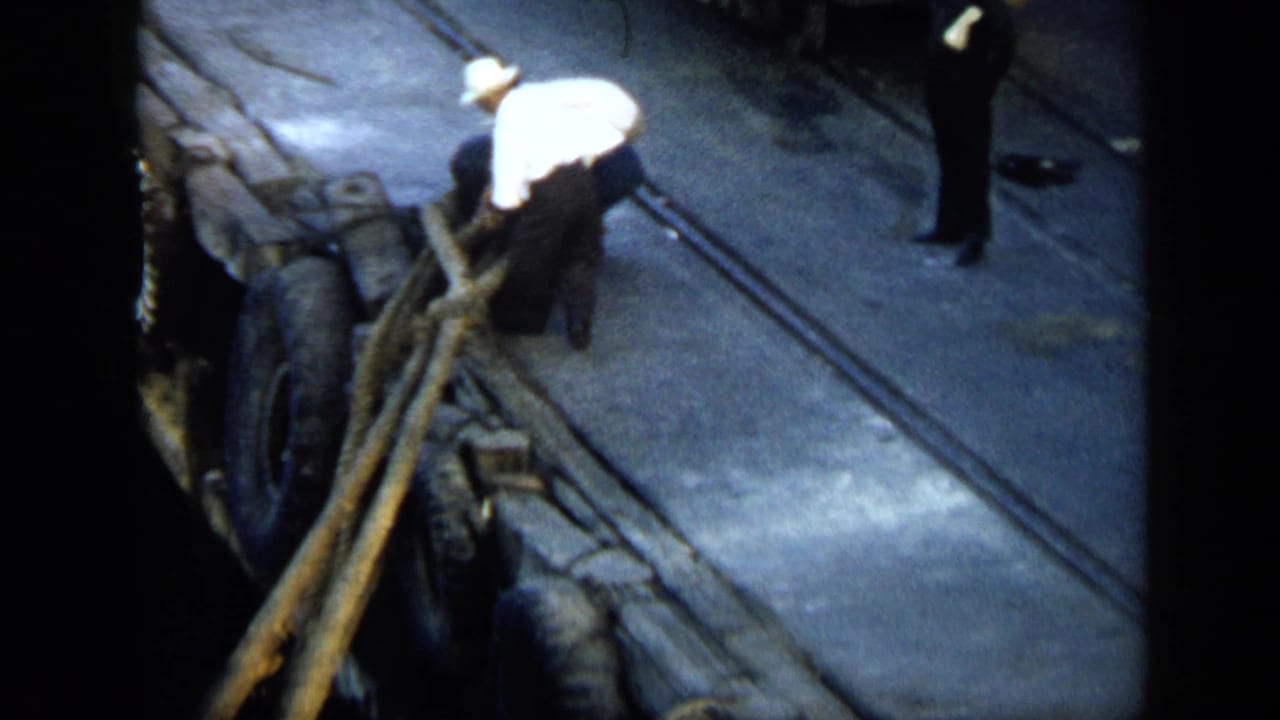 In Venezuela 1959 home-movie footage, a man in a white shirt works with heavy dock lines on a quay as another person stands back.