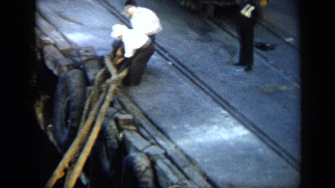 A man in a white shirt leans over heavy ropes on a Venezuelan quay, with a dark-clothed figure standing in the background.