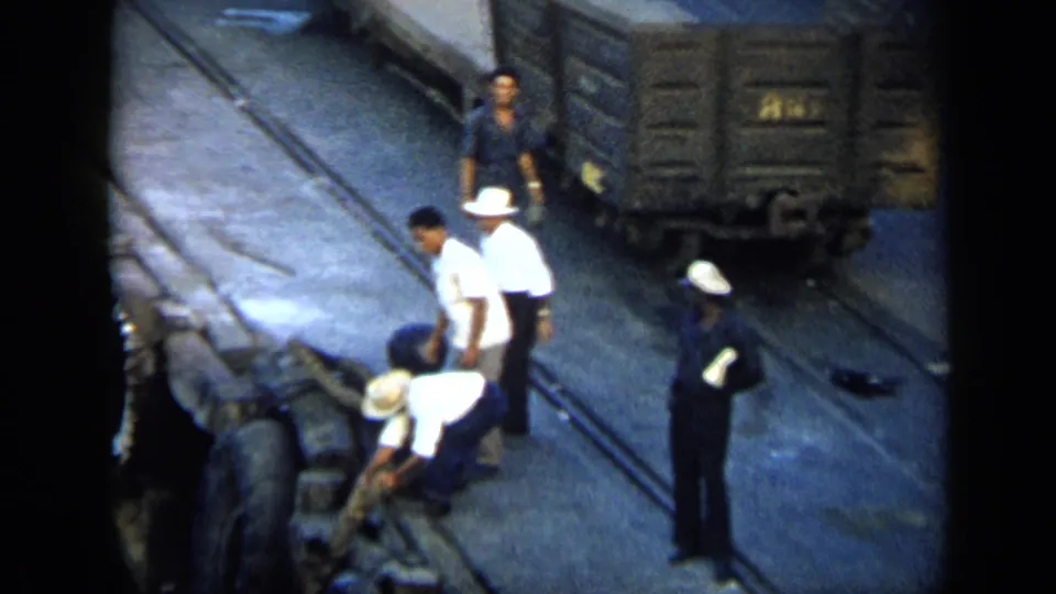 Rail-yard workers in white hats and dark uniforms tend equipment near freight cars in 1959 Venezuela.