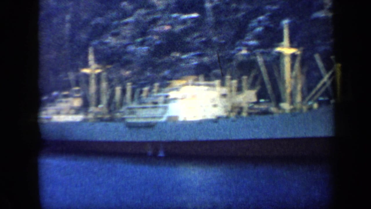 1959 Venezuela harbor view with a large moored ship, masts or cranes, blue water, and a steep tree-covered shore behind it.