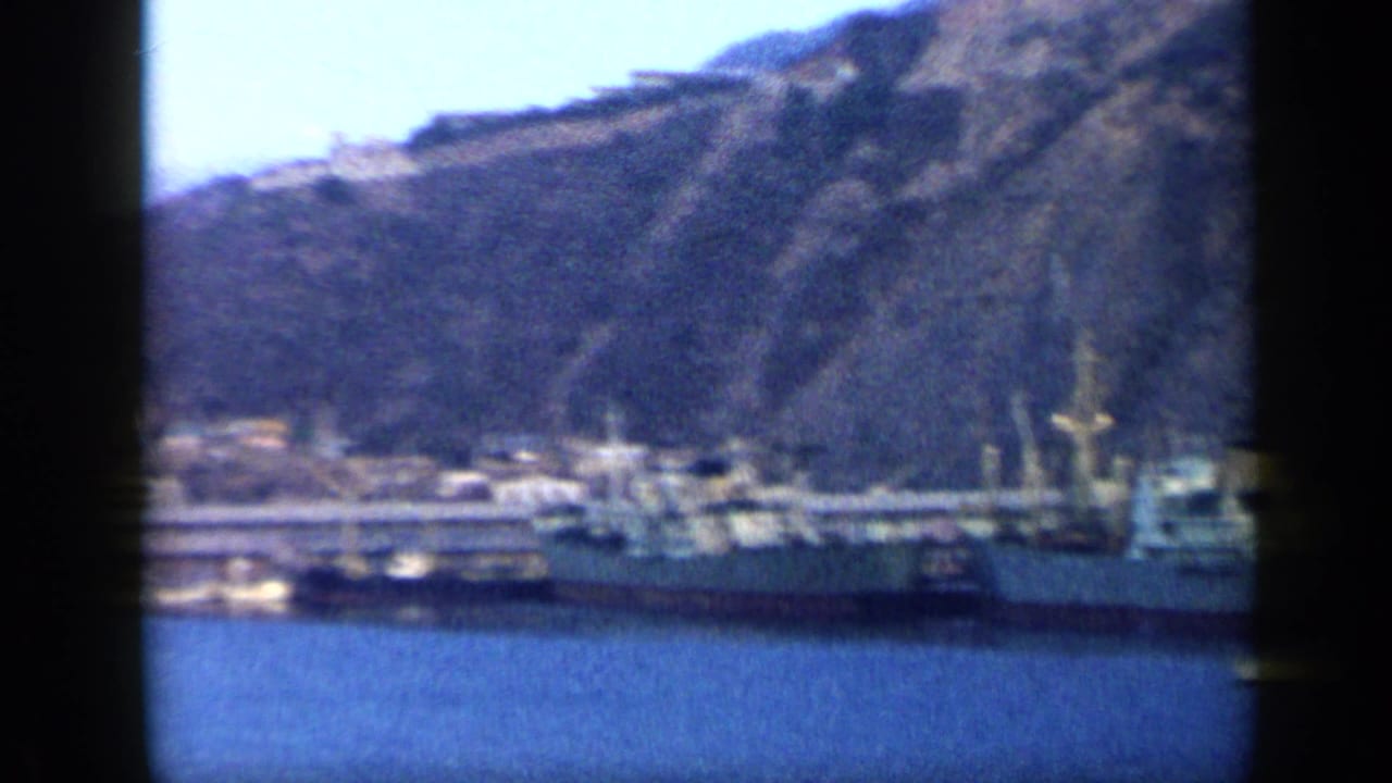 Soft 8mm frame of a Venezuelan waterfront with moored vessels, low dock buildings, blue water, and a steep hillside above the port.