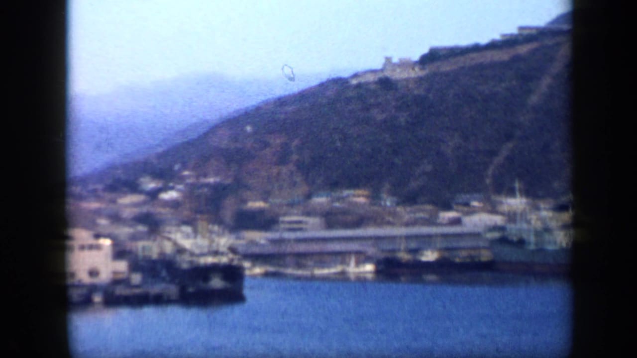 Wide 1959 Venezuela harbor view with ships near the shore, waterfront buildings, and hillside development rising above the water.