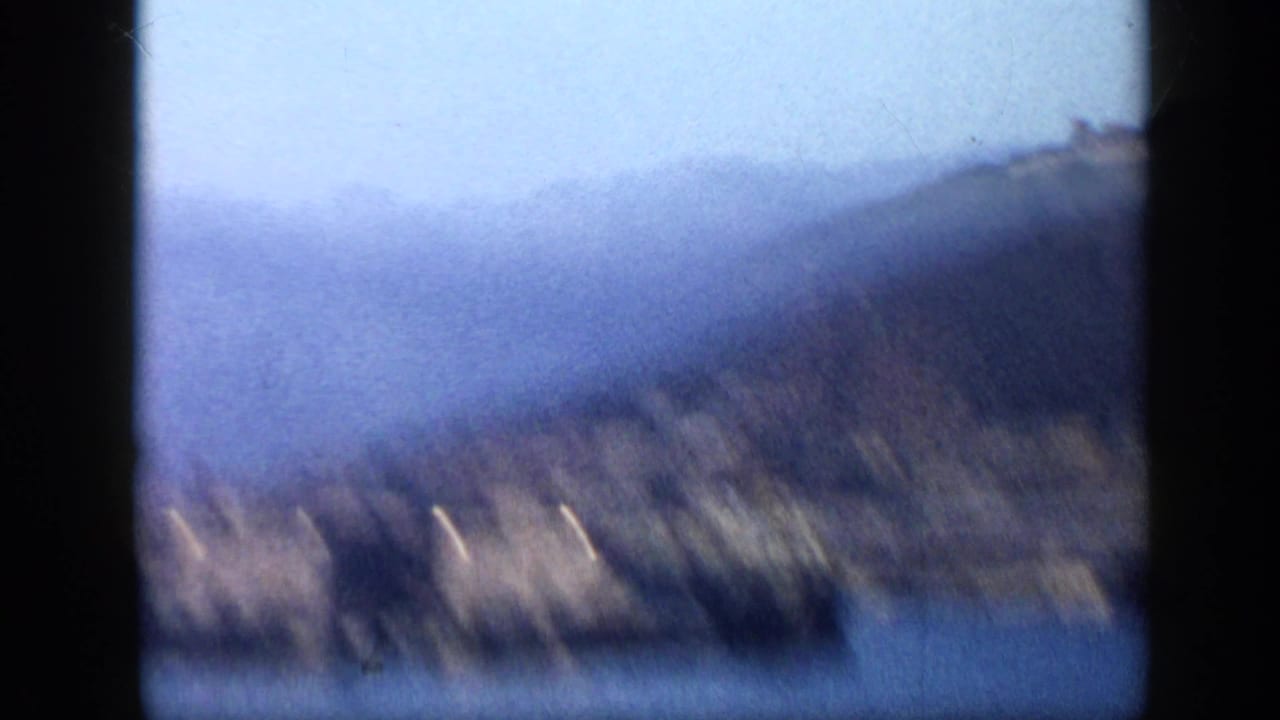 1959 Venezuela home-movie frame showing a hazy hillside coast across blue harbor water, with heavy film gate shadows and soft focus.