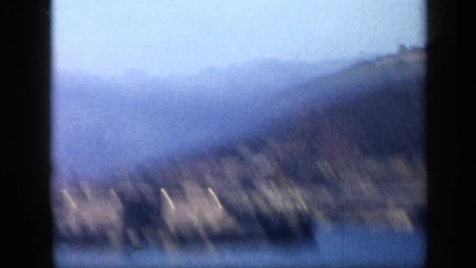 1959 Venezuela home-movie frame showing a hazy hillside coast across blue harbor water, with heavy film gate shadows and soft focus.