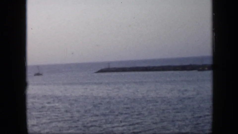 1959 Venezuela coastal harbor view with open water, a long breakwater or jetty, hazy sky, and a small distant vessel near the shoreline.