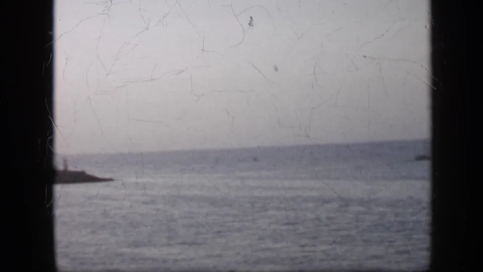 1959 Venezuela 8mm frame with a pale ocean horizon, distant low land, and a dark foreground divider crossing the view.