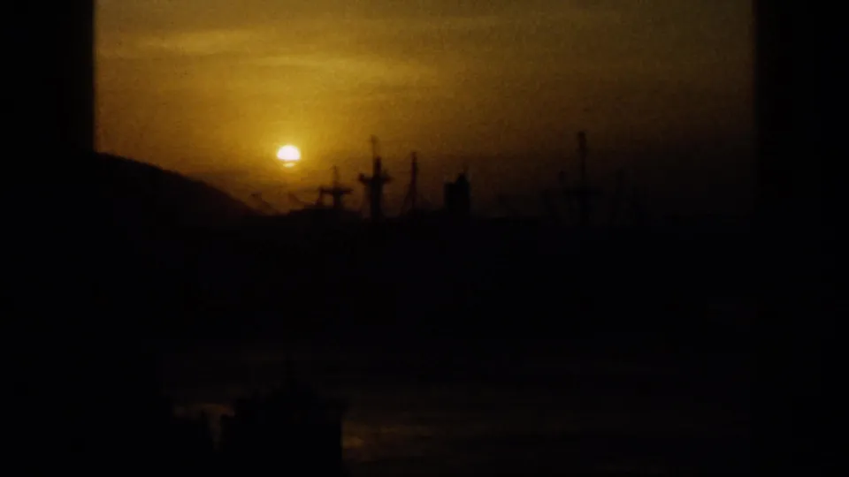 1959 Venezuela 8mm frame showing an orange sun near the horizon, silhouetted harbor structures, water, and low hills.