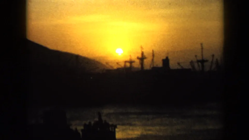 1959 Venezuela 8mm frame with a bright sunset over water, silhouetted cranes or masts, and low hills around a harbor.