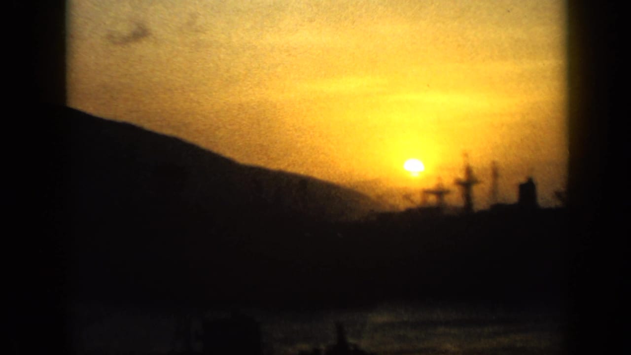 1959 Venezuela home-movie frame showing a low sun over water, dark mountain ridge, and silhouetted harbor structures.