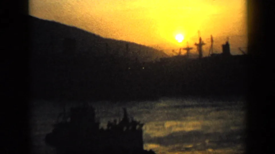 1959 Venezuela dusk view with the sun above a mountain slope, silhouetted cranes, water, and a small boat shape below.