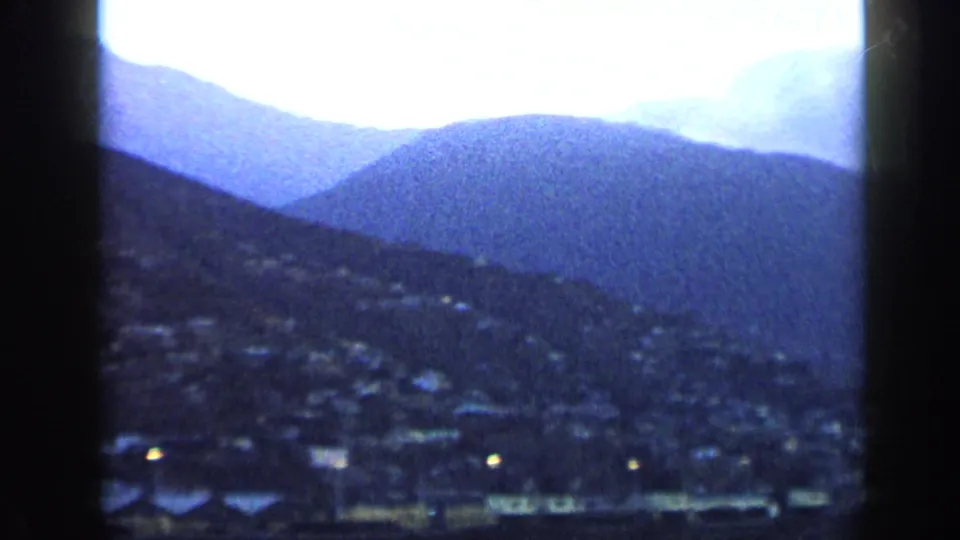 1959 Venezuela frame showing a dusk valley town, blue mountain slopes, faint rooftops, and a few lights in the foreground.