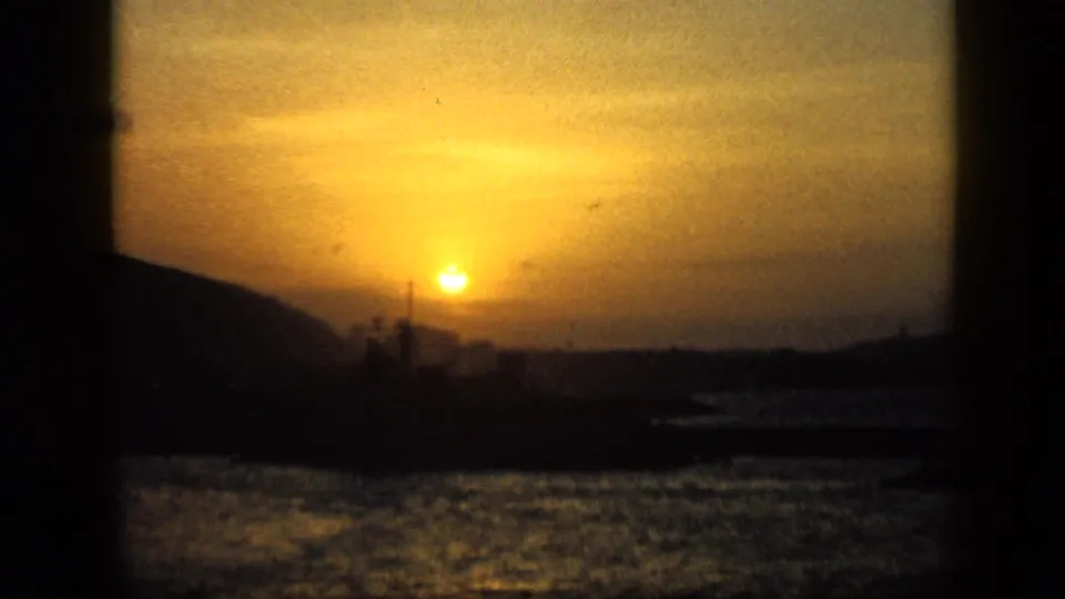 1959 Venezuela 8mm frame of a low golden sun over bay water, with the shoreline and ship forms reduced to silhouettes.
