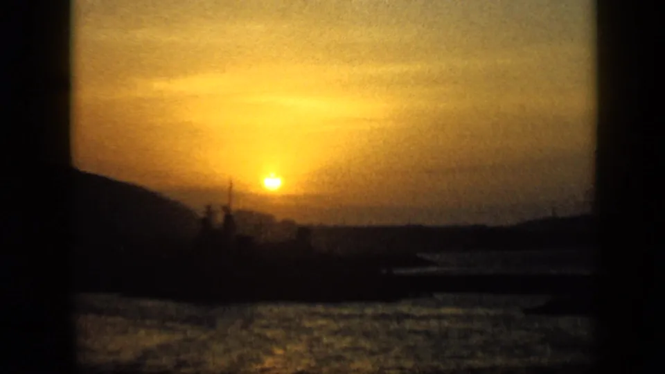 A warm sunset over a Venezuelan bay, with silhouetted shoreline, harbor structures, and water texture on vintage 8mm film.