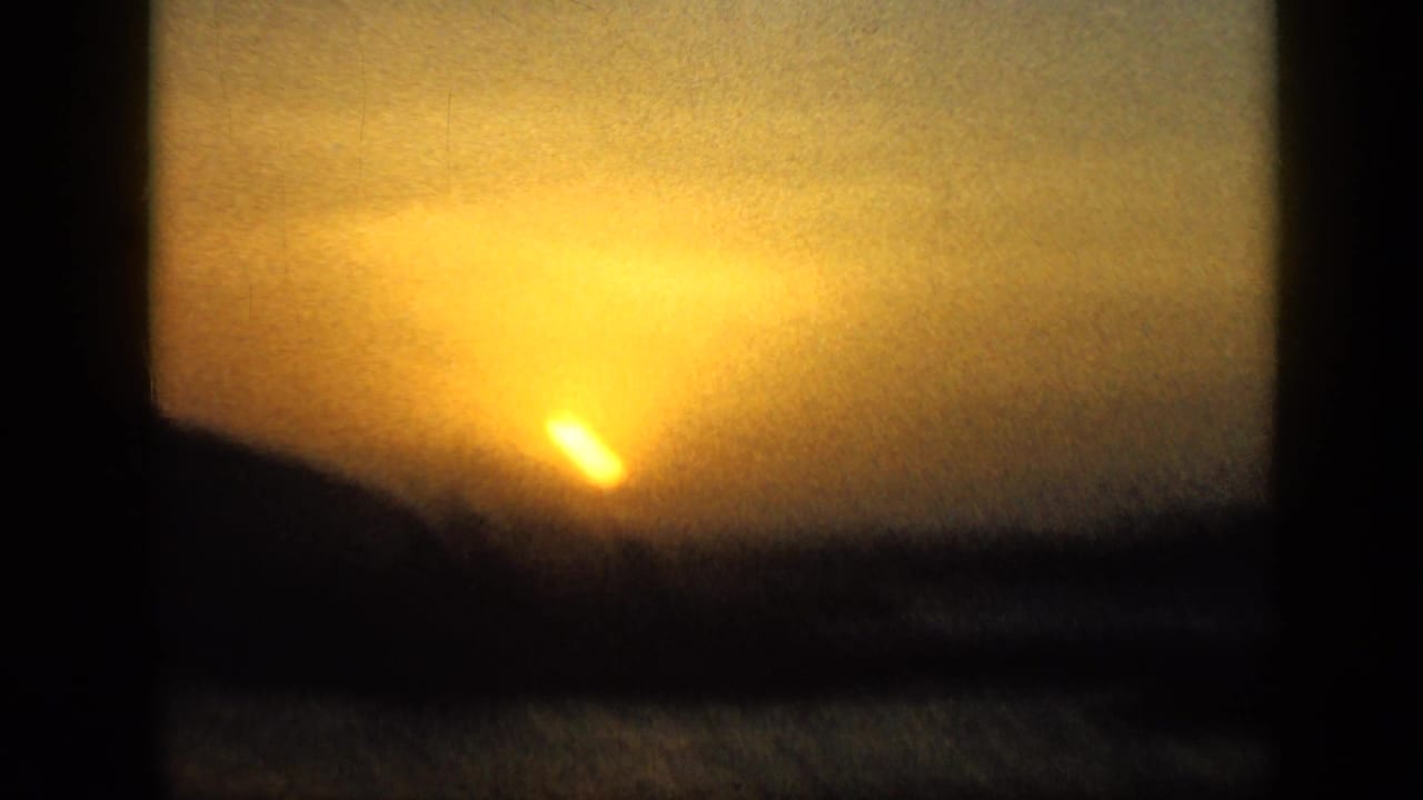 Low sun setting behind dark hills with water in the foreground.
