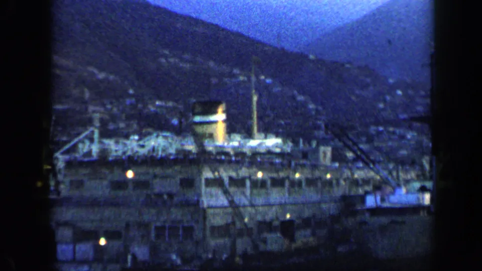 1959 Venezuela port scene with a large docked vessel, harbor equipment, small lights, and hillside buildings in blue twilight.