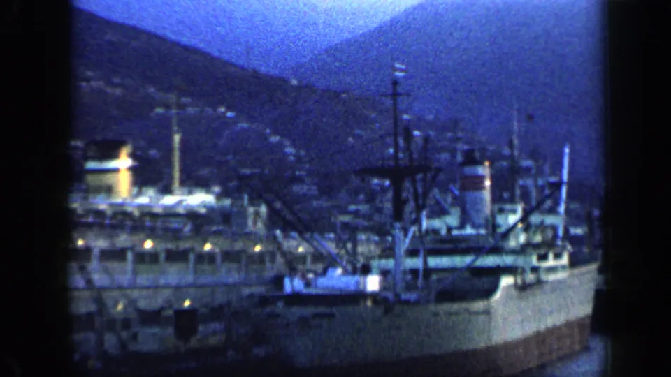 1959 Venezuela harbor frame showing a large ship beside port cranes and masts, backed by dark hills in blue evening light.