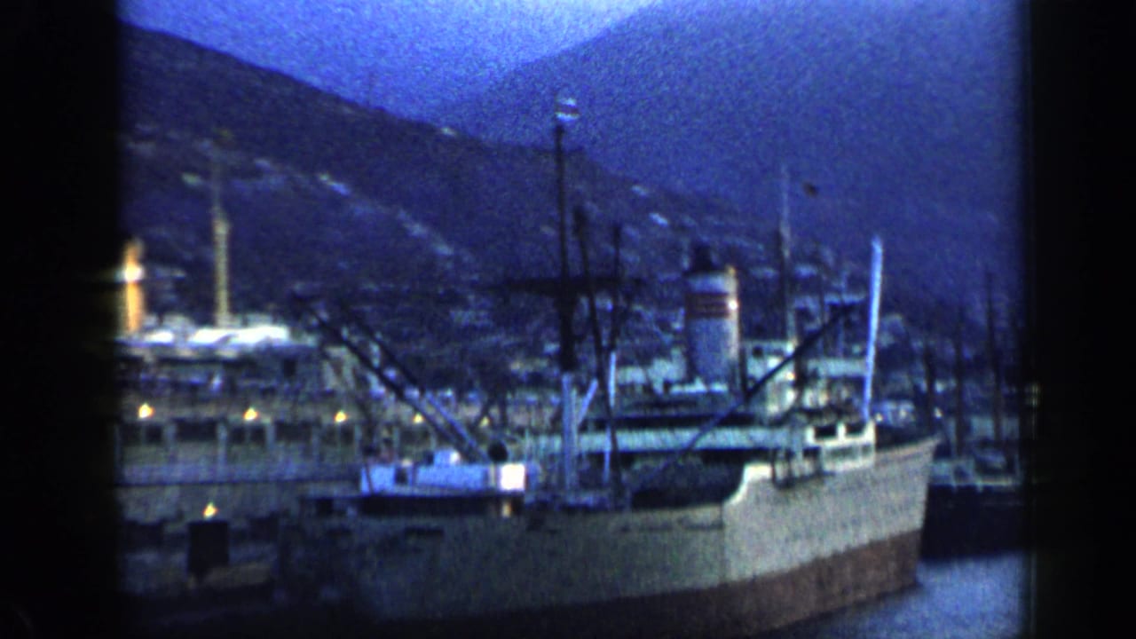 1959 Venezuela 8mm frame showing moored cargo ships, dock structures, harbor water, and a hillside city under blue dusk light.