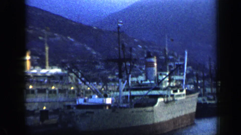 1959 Venezuela 8mm frame showing moored cargo ships, dock structures, harbor water, and a hillside city under blue dusk light.