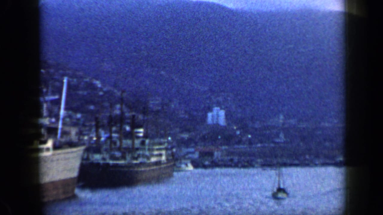 1959 Venezuela 8mm frame showing a wider harbor view with moored boats, blue water, hillside buildings, and mountains in dim evening light.