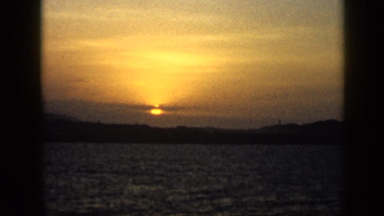 1959 Venezuela 8mm frame showing the sun setting over open water, with a dark coastal horizon and warm orange sky.